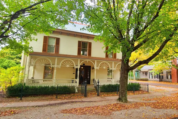 A two-story white historic house with decorative porch arches and brown shutters stands behind a black iron fence, framed by a large leafy tree and a street lined with additional historic buildings.