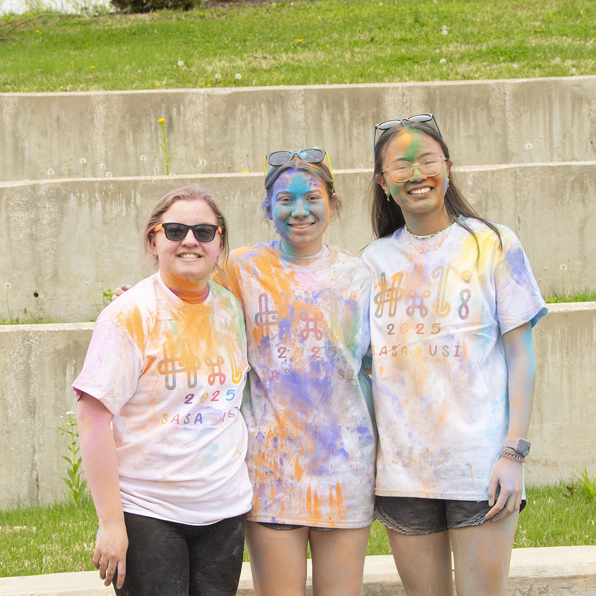 Three students covered in colorful cornflour dust smile for a photo at the Holi Festival at USI.