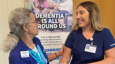 An older woman (Teepa Snow) standing to the side, looking with a smile, and touching the arm of a USI Nursing student.