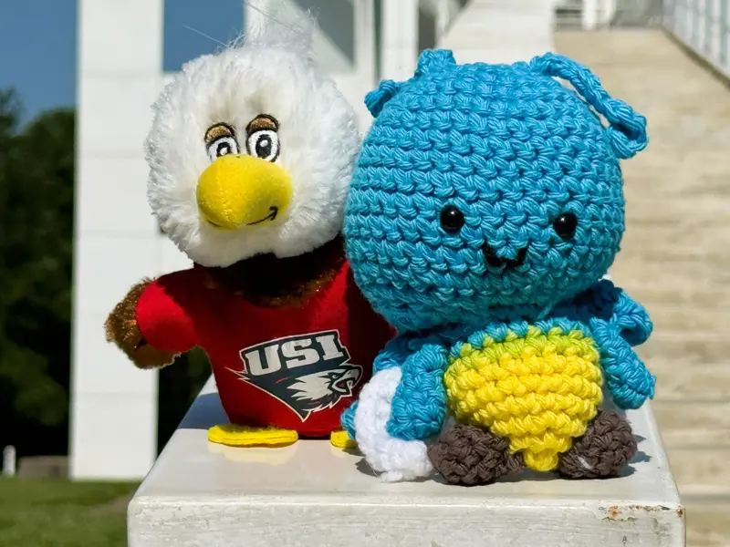 Plush blue crocheted firefly toy beside a USI eagle mascot plush wearing a red shirt.