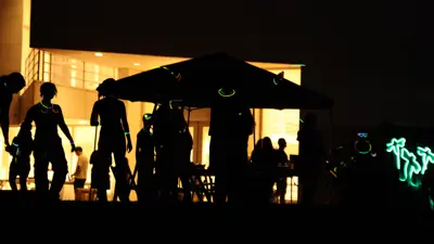 Silhouetted visitors stand and walk beneath a large canopy at night, wearing glowing bracelets and necklaces, with the illuminated Atheneum building visible behind them and neon light shapes glowing in the background.