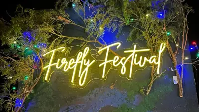 Neon sign reading “Firefly Festival” glowing in warm yellow light, mounted on a dark wall and surrounded by tree branches and multicolored string lights at night.