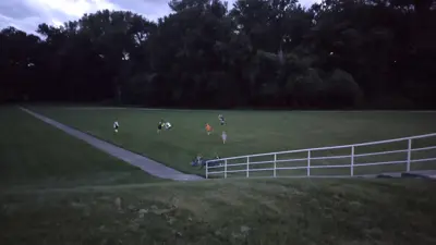 Open grassy field at dusk with a paved walking path and white railing in the foreground. Several small groups of visitors are scattered across the lawn, some with faint glowing lights, with a tree line in the background under a cloudy evening sky.