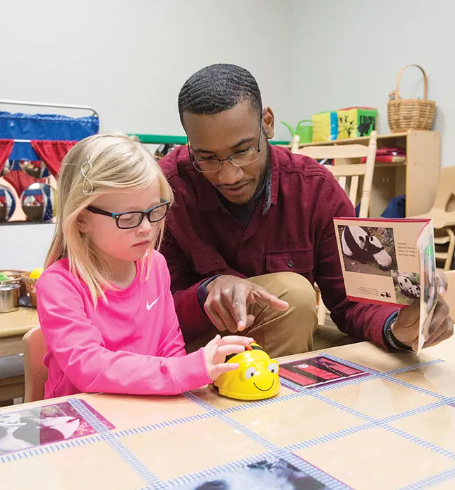 A teacher candidate works with a young student wearing a pink shirt and glasses. The teacher is holding a book with pictures of pandas. The student is playing with a bumble bee toy on the table.