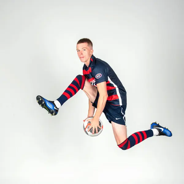 A member of soccer team jumps in the air and passes a soccer ball between his legs like he is in a slam dunk contest for media day.
