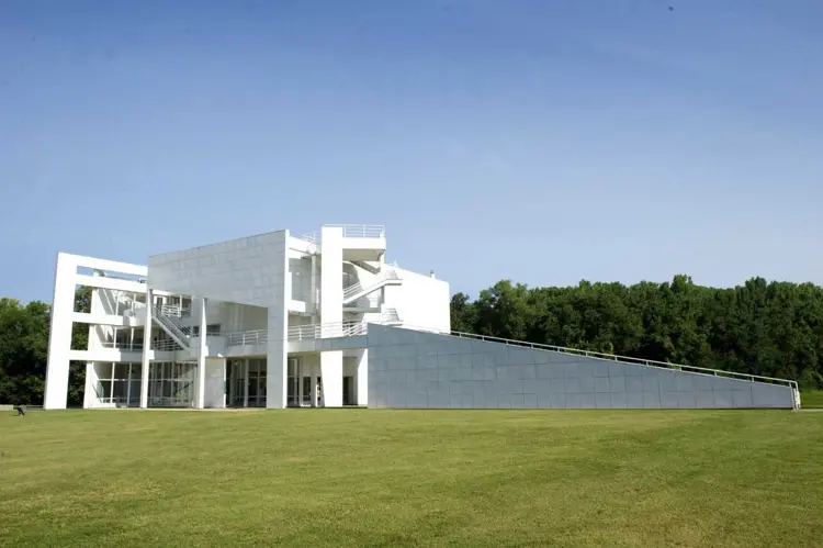 The white modernist Atheneum building, with angular forms and exterior staircases, stands on a wide grassy lawn under a clear blue sky, with a tree line in the background.