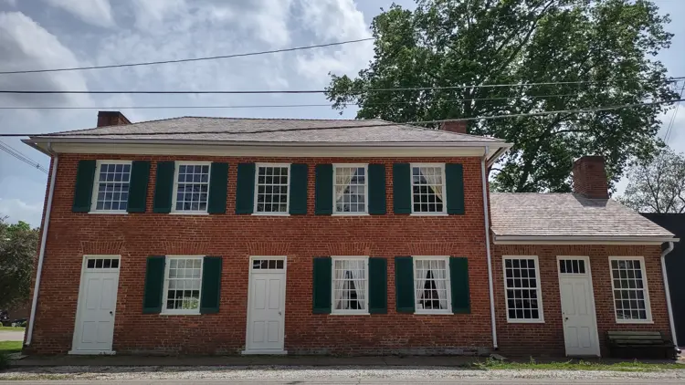 A two-story historic red brick building with green shutters and evenly spaced windows extends along a street, with multiple white doors and chimneys visible behind overhead utility lines.