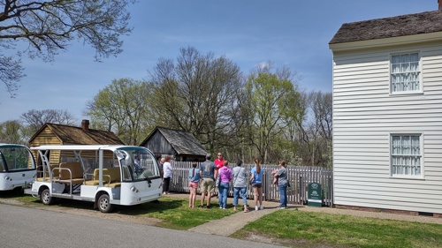 A small group of visitors stands along a white picket fence listening to a guide in a historic outdoor setting. Two open-sided electric tour trams are parked on the left, while a white clapboard house fills the right side of the frame. Behind the group are several small wooden buildings and trees with early spring foliage under a partly cloudy sky.