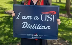 Student in cap and gown holding sign that says "I am a USI Dietitian"