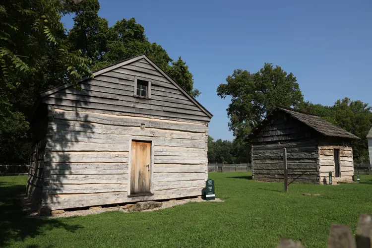 Two small historic log cabins with weathered wood siding stand on a grassy lawn surrounded by trees, with one cabin in the foreground and another slightly behind it, enclosed by a simple fence under a clear blue sky.