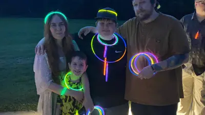 Group of four people standing outdoors at dusk wearing glowing accessories, including neon necklaces and bracelets. The group poses on a paved path with a grassy field and trees behind them, holding glow sticks and rings that emit bright green, blue, and orange light.
